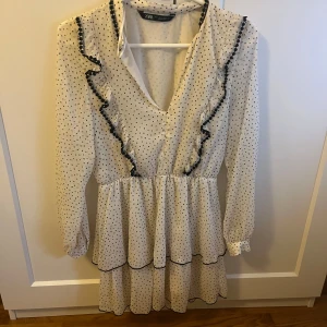 Vit prickig omlottklänning från Zara - Supersöt vit omlottklänning från Zara med svarta prickar och volangdetaljer längs v-ringningen. Klänningen har lång ärm och två volanger i kjolen för extra flow. Perfekt för vår och sommar!