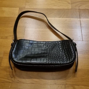 Svart axelväska i krokopräglat skinn från NLY Accessories - Säljer en svart axelväska från NLY Accessories i krokopräglat skinnimitation. Väskan har en stilren form, ett huvudfack med dragkedja och en smal axelrem. Perfekt för att lyfta din outfit med en trendig accessoar.