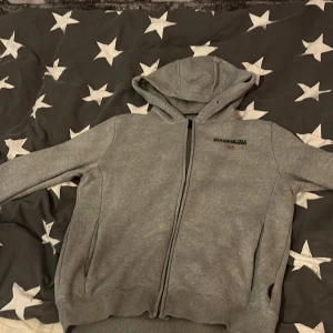 Grå hoodie med dragkedja från Napapijri - Säljer en grå hoodie från Napapijri med dragkedja framtill och huva. Tröjan har logga och norsk flagga broderad på bröstet samt på ryggen. Perfekt för en avslappnad stil och enkel att matcha.