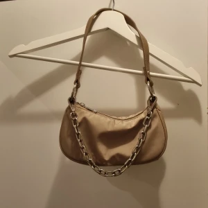 Beige axelväska med kedjedetalj - Säljer en beige axelväska med cool silverfärgad kedja framtill. Väskan har dragkedja upptill och ett stilrent, minimalistiskt utseende. Perfekt för att lyfta din outfit med en trendig accessoar.