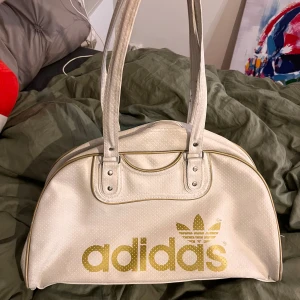Vit axelväska från Adidas med guldlogga - Säljer en vit axelväska från Adidas med stor guldig logga framtill. Väskan har två långa handtag, dragkedja upptill och är tillverkad i syntetmaterial med diskret prickigt mönster. Perfekt för att bära med sig mycket packning! Väskan är lite skitig kan försöka tvätta av den innan postningen, en liten defekt på ena dragkedjan men annars är den perfekt. Mått: Bredd: 15cm Längd: 43cm Djup: 25cm💞
