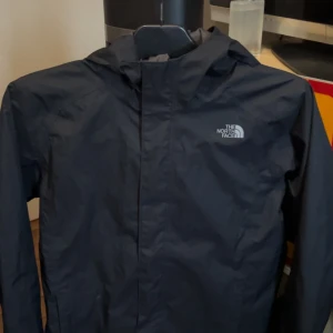 Svart vindjacka från The North Face - Säljer en svart vindjacka från The North Face med huva och dragkedja. Jackan är lätt och har en enkel, stilren design med logga på bröstet och på ryggen. Perfekt för blåsiga dagar och enkel att packa ner i väskan.