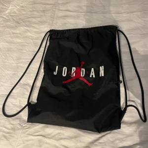 Svart gympåse från Jordan - Säljer en svart gympåse från Jordan med klassisk logga i rött och vitt. Väskan har dragskostängning och en praktisk dragkedjeficka på sidan. Perfekt för träning eller vardagsbruk.
