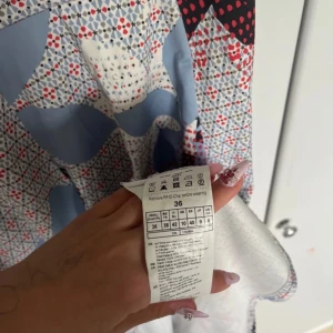 Mönstrad klänning från Taifun - Säljer en äkta klänning från Taifun med geometriskt mönster i blått, rött, svart och vitt. Klänningen har en feminin passform, är ärmlös och har en rund halsringning. Materialet är mjukt och lätt, perfekt för varmare dagar.