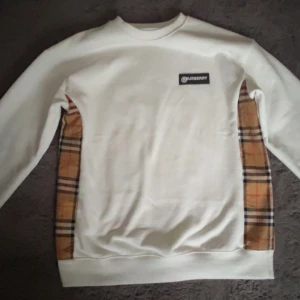 Burberry Tröja - Säljer en vit Tröja från Burberry med klassiska rutiga paneler på sidorna och en svart logotyp-patch på bröstet. Tröjan har rund hals och långa ärmar. Perfekt för dig som gillar stilrena plagg med en twist.