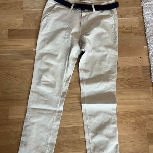 Beige chinos från NN07 med bälte - Snygga beige chinos från NN07 med medföljande mörkblått flätat bälte. Klassisk rak passform med fickor i sidorna och bak. Perfekta för en stilren och avslappnad look.