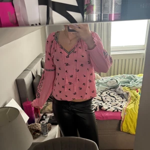 Rosa blus med broderade detaljer - Säljer en söt rosa blus med svarta broderade detaljer och små blommor. Blusen har v-ringning med knyt och långa ärmar med elastiska muddar. Perfekt för dig som gillar boho-stil!