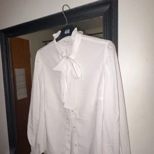 Vit blus med rosett från APT - Säljer en klassisk vit blus från APT med lång ärm och snygg rosettdetalj vid halsen. Blusen har knappar framtill och en elegant, feminin look. Perfekt att styla med jeans eller kjol.