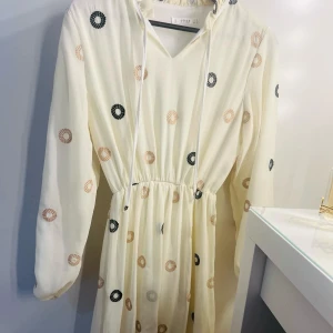 Vit långärmad klänning med broderade cirklar från By Malina - Säljer en vit långärmad klänning från By Malina med broderade cirklar i svart och beige. Klänningen har v-ringning med knytband och resår i midjan för en smickrande passform. Perfekt för vår och sommar! Kan änvändas även för storlek 38