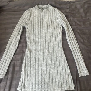 Ljusgrå ribbad långklänning med lång ärm - Säljer en ljusgrå ribbad långklänning med lång ärm och hög hals. Klänningen har en tight passform och är tillverkad i ett mjukt, stretchigt material som sitter snyggt på kroppen. Perfekt för dig som gillar en enkel men stilren look.