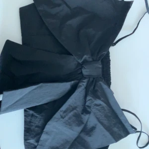 Svart croppad topp med stor rosett från Divided H&M - Säljer en svart croppad topp från Divided H&M med en stor rosett framtill. Toppen har smala axelband och elastisk rygg för en bekväm passform. Perfekt för att sticka ut med en snygg detalj!