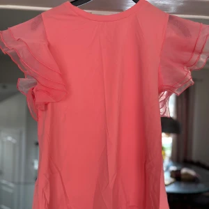 Rosa/orange blus/t-shirt med volangärmar - Säljer en söt rosa blus med volangärmar. Toppen har rund halsringning och dubbla lager volanger på ärmarna som ger en luftig och feminin känsla. Perfekt för dig som vill ha något färgglatt och lekfullt i garderoben!