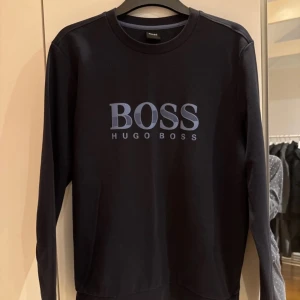 Svart långärmad tröja från Hugo Boss - Säljer en svart långärmad tröja från Hugo Boss med stor BOSS-logga i blått framtill. Tröjan har rund halsringning och en stilren, enkel design som passar perfekt till vardags. Klassisk passform och mjukt material.