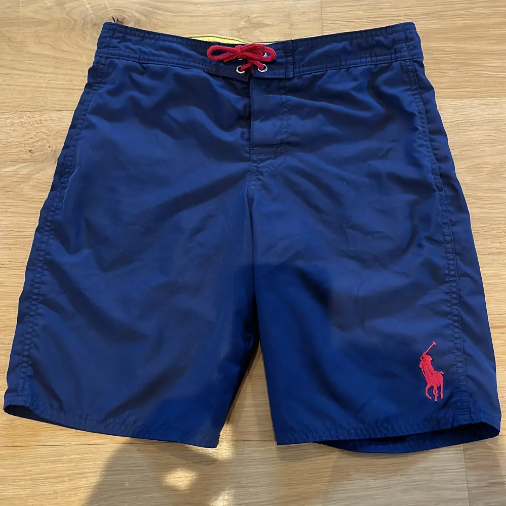 Snygga marinblå badshorts från Polo Ralph Lauren med röd broderad logga på benet och röd snörning i midjan. Klassisk modell med normal passform, perfekt för sommaren. Tillverkade i polyester för snabb torkning. Storlek: M (10-12). Hör av dig vid frågor!. Muu.