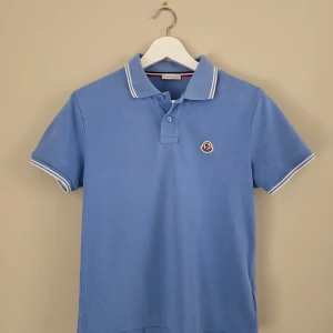 Blå pikétröja från Moncler passar 13-15 år - Snygg blå pikétröja från Moncler med klassisk krage och vita ränder på krage och ärmslut. Tröjan har korta ärmar, knappar framtill och Moncler-logga broderad på bröstet. Markerad som Small men skulle säga att den är liten i storleken. 60 cm från axel nederkant, 42 cm över bröstet på framsidan. Passar personer 150-160 cm lång 