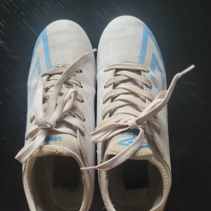 Vita och blå fotbollsskor från Umbro - Säljer ett par vita fotbollsskor från Umbro med blå detaljer och klassisk design. Skorna har snörning och är gjorda i syntetmaterial, perfekta för spel på konstgräs eller gräsplan. Snygg och sportig look med tydliga Umbro-loggor på sidorna.