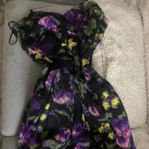 Svart mini klänning med blommigt mönster - Säljer en svart mini klänning med färgstarkt blommigt mönster i lila, grönt och gult. Klänningen har en markerad midja och lätt utsvängd kjol, perfekt för dig som vill sticka ut.