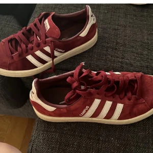 Vinröda Adidas Campus sneakers - Säljer ett par vinröda Adidas Campus sneakers med vita ränder och vit sula. Skorna har klassisk snörning och är gjorda i mocka med detaljer i syntet. Perfekta för dig som gillar retrostil och vill ha något som sticker ut.