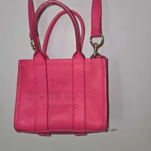 Rosa handväska från Marc Jacobs - Säljer en snygg rosa handväska från Marc Jacobs med präglad text 'The Tote Bag' på framsidan. Väskan har både handtag och en justerbar axelrem i samma färg. Tillverkad i skinn med struktur och guldiga metalldetaljer. 300