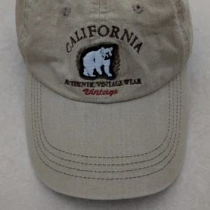 Beige keps med broderad björn och text - Snygg beige keps med broderad vit björn och texten 'California' samt 'Authentic Vintage Wear Vintage' framtill. Justerbar rem baktill för perfekt passform. Perfekt accessoar för en avslappnad stil. Kepsen har aldrig använts 