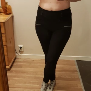 Svarta thigts byxor med fickor strl M  - Svarta leggings med hög midja och dekorativa dragkedjor framtill. Byxorna har knappar vid midjan och är tillverkade i stretchigt material för en tight passform. Perfekta för en stilren och bekväm look. Ricktiga fickor