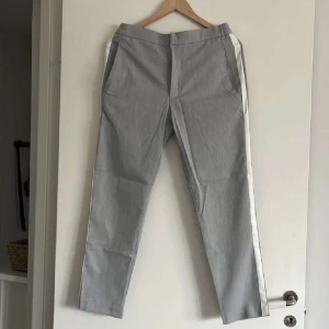Grå byxor med vit sidoremsa - Snygga grå byxor med vit sidoremsa och elastisk midja. Byxorna har raka ben och fickor fram samt en bakficka. Perfekta för en stilren och sportig look.