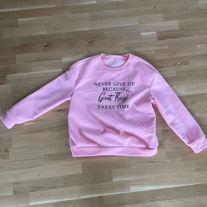 Rosa sweatshirt med text - Säljer en ljusrosa sweatshirt med svart text på bröstet: 'Never give up because great things take time' och ett litet hjärta. Tröjan har rund hals och långa ärmar. Mjuk insida i fleece för extra komfort.