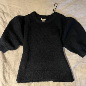 Svart stickad tröja med puffärmar från Gina Tricot - Säljer en svart stickad tröja från Gina Tricot med trendiga puffärmar och rund halsringning. Tröjan har en klassisk passform och är perfekt för lager-på-lager-stil. Superenkel att matcha med både jeans och kjol.