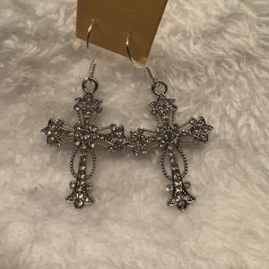 Silvriga korsörhängen med glittriga stenar - Säljer ett par snygga örhängen i form av kors, dekorerade med små glittriga stenar. Perfekta för dig som vill sticka ut med en cool accessoar. Hängande modell i silverfärg. Krokarna är i sterling silver 