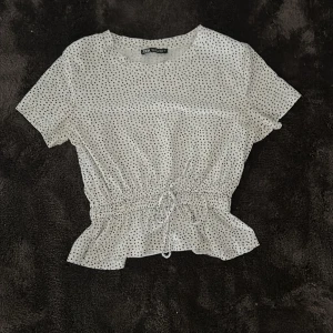 Vit prickig blus från Zara - Säljer en söt vit blus från Zara med svarta prickar. Toppen har korta ärmar, rund halsringning och knyt i midjan som ger en fin volangkant nedtill. Perfekt för en trendig och avslappnad look.