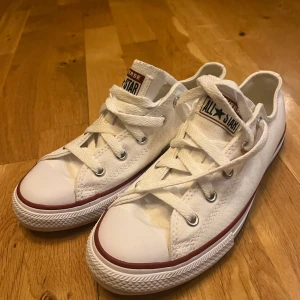 Vita Converse All Star sneakers - Klassiska vita Converse All Star sneakers med lågt skaft och snörning. Skorna har vit canvas, gummisula och röda detaljer längs sulkanten. Perfekta för en avslappnad och stilren look.