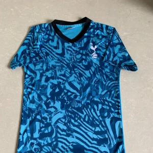 Blå Tottenham Hotspur fotbollströja - Säljer en blå fotbollströja från Tottenham Hotspur med svart V-ringad krage och abstrakt mönster i olika blå nyanser. Tröjan har klubbens emblem på bröstet och är tillverkad i ett lätt och ventilerande material. Perfekt för fotbollsträning eller match.