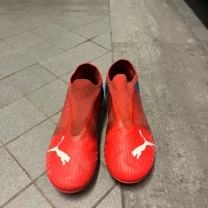 Röda och blå fotbollsskor från Puma - Säljer ett par röda och blå fotbollsskor från Puma med vit logga på ovansidan och texten 'ULTRA' på sidan. Skorna har en strömlinjeformad design utan snörning och är tillverkade i syntetmaterial för bästa prestanda på planen. Nytt pris 1100 typ Mitt pris 400