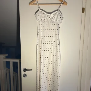 Vit långklänning med svarta prickar - Supersöt långklänning i vit satin med svarta prickar och smala axelband. Klänningen har en feminin passform med markerad byst och är perfekt för sommarens alla tillfällen. Snygg och tidlös design!