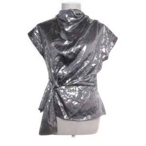 Zara blus med knyt - Säljer en glansig silverfärgad blus med korta ärmar och hög krage. Blusen har ett knyt i midjan och ett diskret mönster i tyget som ger en lyxig känsla. Perfekt för dig som vill sticka ut med en unik topp!