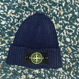  Stone Island mössa - Mörkblå stickad mössa från Stone Island med klassisk broderad logotyp framtill. Mössan är ribbstickad och har en uppvikt kant. Perfekt för kyliga dagar och snygg till streetwear-stilen. One size. Helt oanvänd 