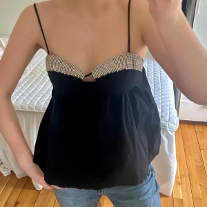 Svart topp med strassdetaljer - Säljer en svart topp med tunna axelband och glittriga strassdetaljer. Superpopulär från Zara. Den är en storlek S men lite långa band så de har jag sytt upp. Saknas några stenar (syns absolut inte). Perfekt till fest!!😍 