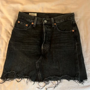 Svart jeanskjol från Levi's Premium - Säljer en svart jeanskjol från Levi's Premium med rå, fransig kant nertill och klassiska fem fickor. Kjolen har knapp- och dragkedjestängning framtill och är i en kort modell. Fint skick 