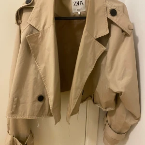 Beige croppad trenchcoat från Zara - Säljer en beige croppad trenchcoat från Zara med stora svarta knappar och bältdetaljer vid ärmsluten. Jackan har klassisk krage och oversized passform, perfekt för en trendig look. Bältet finns även med.