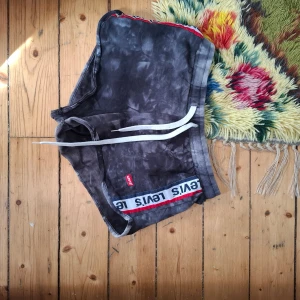 Svarta shorts från Levi's med batikmönster - Säljer ett par svarta shorts från Levi's med batikmönster och vit snörning i midjan. Shortsen har en vit och röd logotypremsa längs kanten och är perfekta för en avslappnad stil.