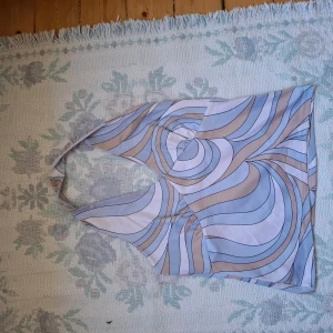 Mönstrad halterneck topp i blått och beige - Säljer en snygg halterneck topp med vågigt mönster i ljusblått, beige och vitt. Toppen har en djup v-ringning och är ärmlös, perfekt för varma dagar eller festival. Materialet känns mjukt och följsamt.