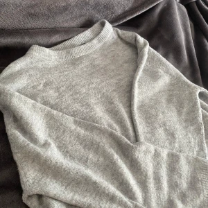 Ljusgrå stickad tröja från 157 - Mysig ljusgrå stickad tröja från 157 med rund halsringning och långa ärmar. Tröjan har ribbade muddar vid hals, ärmslut och nederkant. Perfekt för lager-på-lager och enkel att matcha med olika outfits.