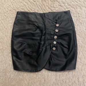 Svart kjol i skinnimitation från NLY Trend - Säljer en svart kort kjol i skinnimitation från NLY Trend. Kjolen har dekorativa knappar framtill med glittriga detaljer och veckad design. Snygg och trendig modell med omlottkänsla och markerad midja.