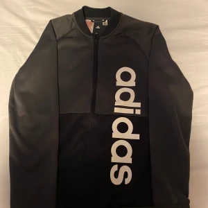 Svart träningsjacka från Adidas - Säljer en svart träningsjacka från Adidas med vit logga längs framsidan. Jackan har dragkedja och lång ärm, perfekt för träning eller sportiga outfits. Enkel och stilren design med klassisk Adidas-känsla.