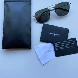 Saint Laurent solglasögon med silverfärgad metallbåge - Nypris  3190kr. Inköpta på synsam. Aldrig använda. Snygga solglasögon från Saint Laurent med rektangulära svarta glas och tunn silverfärgad metallbåge. Klassisk och stilren design med svarta skalmar. Perfekt accessoar för att lyfta din look.