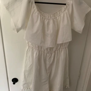 Vit offshoulder byxdress med volang - Superfin vit byxdrss med offshoulder och volang upptill. Klänningen har resår i midjan och axlarna för en bekväm passform. Perfekt för sommarens alla tillfällen!