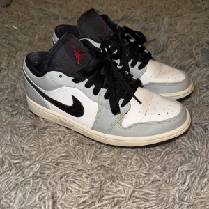 Nike Air Jordan 1 Smoke Grey Low - Säljer ett par Nike Air Jordan 1 Smoke Grey Low, vit och grå färgkombination. Skorna har klassisk svart swoosh, röd logga på hälen och svart snörning. Perfekta för dig som gillar streetwear och sneakers med ikonisk design.