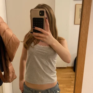 Grå linnetopp med spets från Gina Tricot - Säljer en ljusgrå linnetopp från Gina Tricot med smala axelband och spetsdetalj vid urringningen. Toppen har en liten rosett framtill och texten 'J'adore' tryckt på bröstet. Perfekt för sommaren!