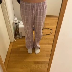 Ljuslila rutiga pyjamasbyxor från Gina Tricot - Mysiga ljuslila pyjamasbyxor från Gina Tricot med vitt rutmönster. Byxorna har resår i midjan och snörning framtill för extra komfort. Perfekta för hemmakvällar eller när du vill chilla.