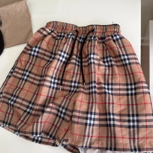 Beige rutiga shorts från Burberry - Snygga beige shorts från Burberry med klassiskt rutigt mönster i svart, vitt och rött. De har elastisk midja med svart snörning och innerfoder i mesh. Perfekta för sommaren och ger en stilren look.